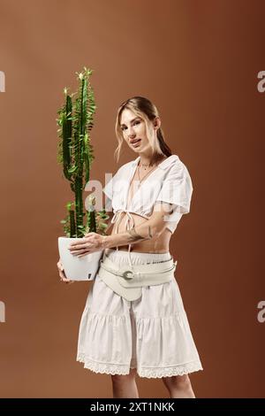 Une jeune femme dans une tenue d'été blanche tient un cactus en pot sur un fond beige. Banque D'Images