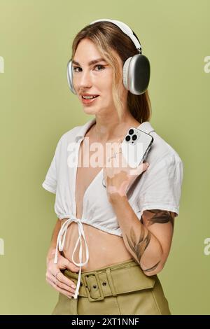 Jeune femme pose en toute confiance dans une tenue blanche sur fond vert, écouteurs sportifs et téléphone de maintien. Banque D'Images