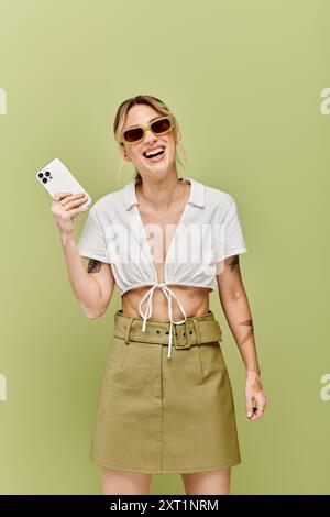 Une jeune femme avec du vitiligo pose dans une tenue d'été blanche sur un fond vert. Elle sourit et tient un téléphone, rayonnant de confiance. Banque D'Images