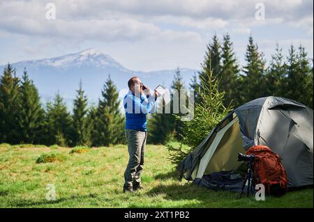 Homme touriste se tient à côté de la tente, parlant au téléphone et tenant le panneau solaire portable. Sac à dos orange et trépied pour appareil photo à proximité. Montagnes enneigées et arbres à feuilles persistantes luxuriantes sous un ciel partiellement nuageux. Banque D'Images