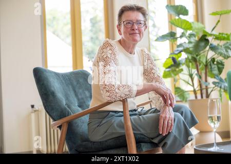 Une femme âgée souriante s'assoit confortablement dans une chaise élégante dans une pièce lumineuse avec des plantes vertes en arrière-plan. Panc05744 Copyright : xConnectxImages Banque D'Images