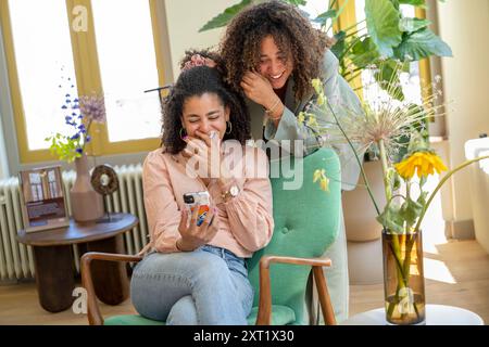 Deux femmes partagent un moment joyeux en regardant ensemble un smartphone entouré de plantes d’intérieur. Panc05754 Copyright : xConnectxImagesx Banque D'Images