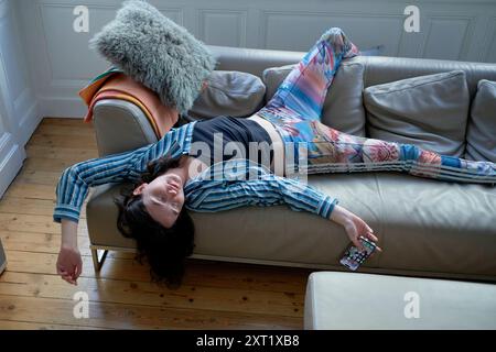 Femme souriante allongée sur un canapé avec des leggings colorés et un dessus rayé, tenant une télécommande. Bola02623 Copyright : xConnectxImagesx Banque D'Images