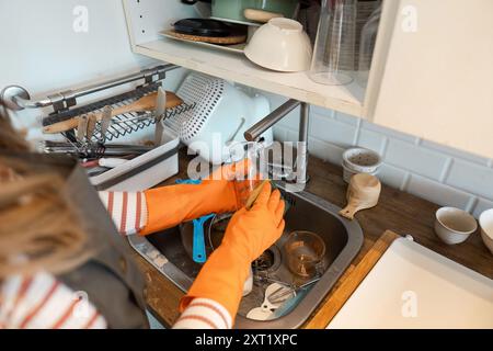 Personne portant des gants orange lavant la vaisselle dans un évier de cuisine moderne avec divers ustensiles et vaisselle Banque D'Images