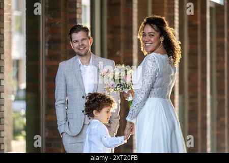 Une famille heureuse habillée en tenue formelle, avec un couple souriant se tenant la main avec leur jeune enfant, posant pour un portrait dans un cadre extérieur ensoleillé Banque D'Images