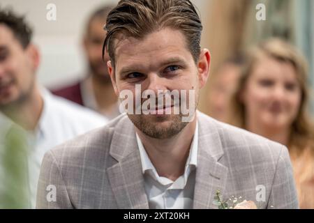 Un marié dans un costume gris à carreaux avec une boutonnière sourit à une cérémonie de mariage, avec des invités flous en arrière-plan. Panc05786 Copyright : xConnect Banque D'Images