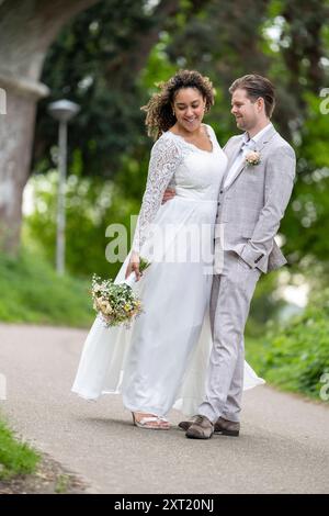 Couple de jeunes mariés marchant main dans la main dans un jardin luxuriant, à la fois souriant et habillé dans une tenue de mariage élégante, avec la mariée tenant un bouquet. panc05794 Banque D'Images