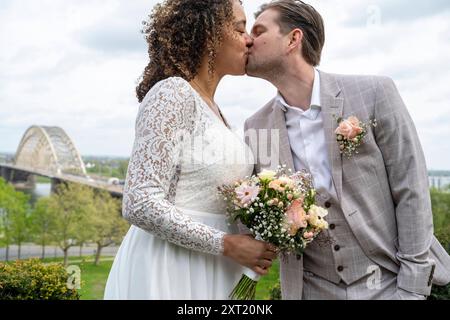 Couple interracial dans la tenue de mariage partageant un baiser à l'extérieur avec un pont en arrière-plan. Panc05801 Copyright : xConnectxImagesx Banque D'Images