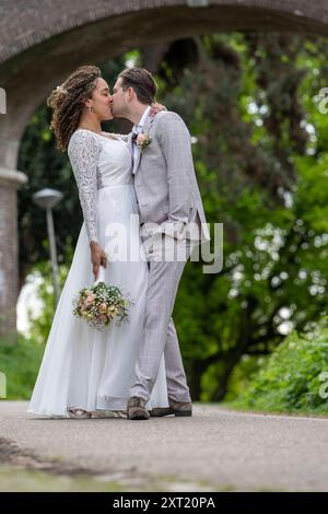 Mariée et mariée partagent un baiser sur un chemin sous un pont, orné d'une élégante tenue de mariage avec un bouquet à la main. Panc05796 Copyright : xConnectxImag Banque D'Images