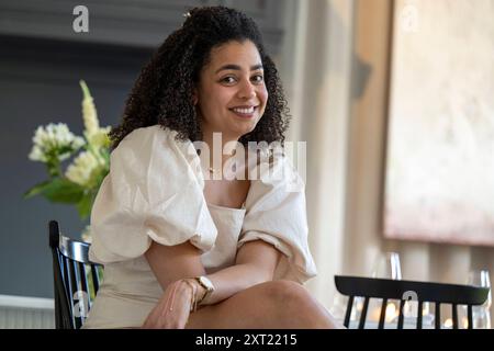 Femme souriante avec les cheveux bouclés assis dans une chaise avec les bras croisés devant une fenêtre, exsudant confiance et confort. Panc05971 Copyright : xConnect Banque D'Images