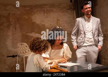 Un marié souriant se tient debout comme une mariée signe un document à une table avec un jeune enfant regardant attentivement dans une pièce au décor rustique. Panc06048 Copyright Banque D'Images