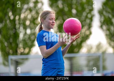 Jeune joueuse de football dans un maillot bleu faisant tourner un ballon de football rose sur le bout de son doigt avec des arbres et un poteau de but en arrière-plan. Panc06282 Copyri Banque D'Images