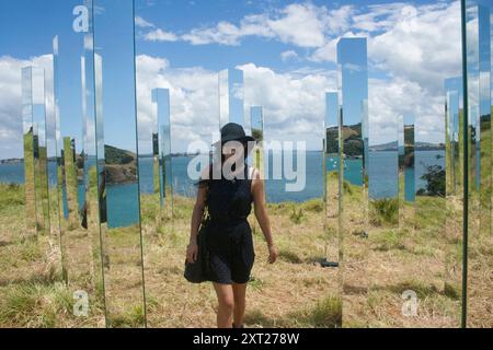 Femme dans une robe noire et un chapeau debout parmi des panneaux verticaux réfléchissants avec une toile de fond côtière pittoresque, Waiheke Island, Nouvelle-Zélande Waiheke Island ne Banque D'Images