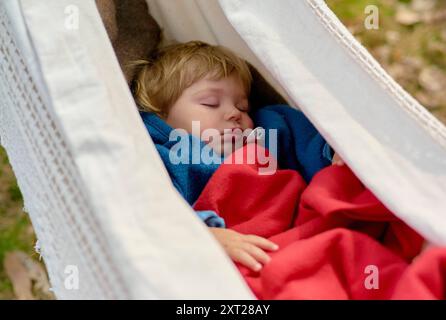 Un jeune enfant dort paisiblement dans un hamac blanc, enveloppé dans une couverture bleue confortable. Bola02900 Copyright : xConnectxImagesx Banque D'Images