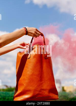 Une main de personne tenant un sac fourre-tout en cuir orange contre une toile de fond d'un ciel bleu avec un nuage de fumée rose. Bola02906 Copyright : xConnectxImagesx RE Banque D'Images