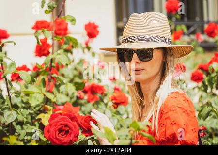 Blonde jeune femme caucasienne en lunettes de soleil et chapeau de paille entourée de roses rouges Banque D'Images