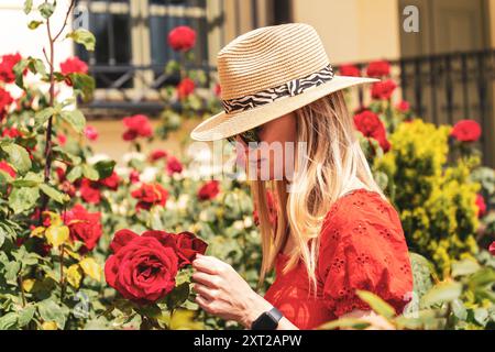 Blonde jeune femme caucasienne en lunettes de soleil et chapeau de paille entourée de roses rouges Banque D'Images
