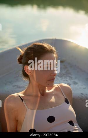 Femme se relaxant dans un bateau se prélassant dans la lumière dorée du soleil pendant le coucher du soleil, portant une robe à pois. Bola03017 Copyright : xConnectxImagesx Banque D'Images