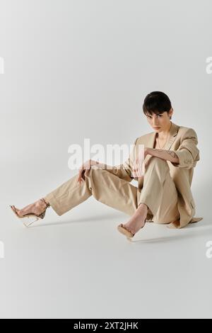 Une femme portant un costume beige et des talons clairs pose avec ses jambes croisées sur un fond blanc. Banque D'Images