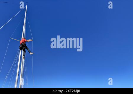 Homme travaillant sur le mât d'un yacht à voile, grimpant avec la corde et la chaise de bosun sur une journée ensoleillée avec le ciel bleu, réparant, fixant les enveloppes de mât Banque D'Images