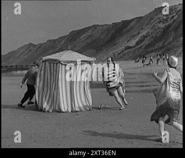 Un acteur masculin avec une moustache et un chapeau Bowler poursuivi autour d'une tente changeante sur une plage par un homme avec un pistolet portant une robe de chambre rayée et un maillot de bain. Un acteur féminin dans Evening Dress Looks on, années 1920 '...Elstree [studios de cinéma] à l'époque des silences...quand le temps était beau bien sûr, au soleil nous sommes allés. Ça... était censé être drôle...'. De "Time to Remember - came the Dawn", 1925 (bobine 1) ; aperçu de l'industrie cinématographique britannique des années 1920 - extraits de grands films muets et de nouvelles. Banque D'Images