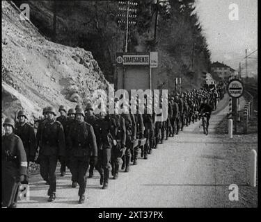 Une ligne de soldats allemands marchant à travers un point de douane le long d'une haute banque avec des arbres à la frontière entre l'Allemagne et l'Autriche avec des panneaux indiquant 'STRAASGRENZE' et 'ZOLL DUANE' de chaque côté de la route et un officiel sur un vélo, 1938. De "Time to Remember - Wind Up week", 1938 (bobine 1) ; film documentaire sur 1938 - les gens prennent conscience de la menace croissante de la guerre. Banque D'Images