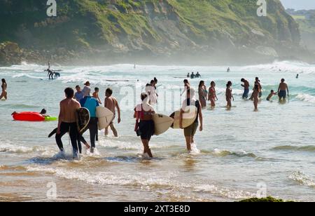 Surfeurs marchant dans le surf portant des planches de surf le long de la plage des surfeurs Playa de los Locos sur un chaud jour de juillet d'été Suances Cantabria Espagne Europe Banque D'Images