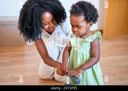 Une joyeuse mère afro-américaine aide sa jeune fille à faire des pas dans une pièce très éclairée, soulignant un moment familial tendre Banque D'Images