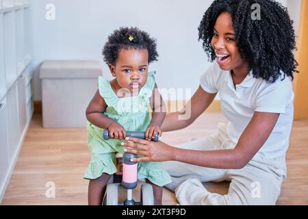 Mère afro-américaine riant avec sa fille en bas âge qui est assise sur un jouet à cheval dans un cadre lumineux, mettant en valeur un moment familial chaleureux Banque D'Images