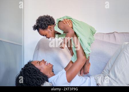 Une mère afro-américaine soulève sa fille dans les airs, souriant joyeusement dans un cadre de maison léger et confortable, mettant en vedette une maman familiale chaleureuse Banque D'Images