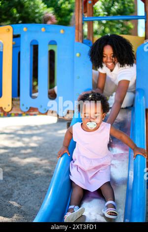 La mère afro-américaine assiste joyeusement sa fille tout-petit comme elle glisse vers le bas d'un terrain de jeu bleu vif diapositive parfaite capture d'un insouciant et lovi Banque D'Images