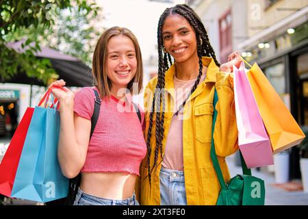 Deux joyeuses amies féminines multiethniques profitant d'une virée shopping dans un cadre urbain, transportant des sacs colorés et regardant l'appareil photo avec smil lumineux Banque D'Images