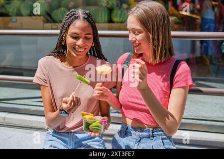 Deux amis partageant un moment joyeux en mangeant des fruits frais par une journée ensoleillée. Ils sont souriants, reflétant une atmosphère chaleureuse et conviviale. Banque D'Images