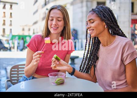 Deux amies, une femme caucasienne et une femme d’ascendance africaine, partagent un moment joyeux en mangeant des fruits dans un bol à une table en plein air dans un setti urbain Banque D'Images