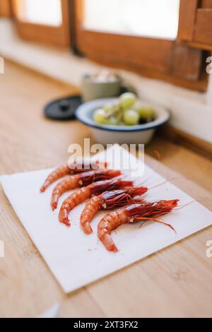 Des crevettes fraîches et vibrantes alignées sur une planche à découper blanche. Dans le fond légèrement concentré, une table en bois contient des plats supplémentaires, y compris un bol de Banque D'Images