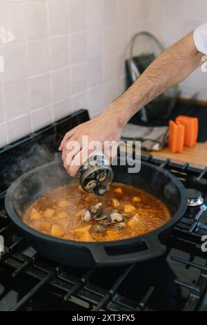 Image recadrée méconnaissable de la main d'un chef masculin assaisonnant un ragoût mijotant avec des moules et des légumes dans une cuisine. De la vapeur s'échappe de la casserole, comme illustré à la Banque D'Images