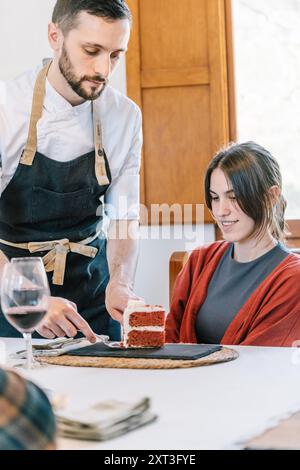 Un chef privé, concentré et regardant vers le bas, sert une tranche de gâteau de velours rouge à une jeune femme à une table à manger. La femme, portant un cardigan rouge, loo Banque D'Images