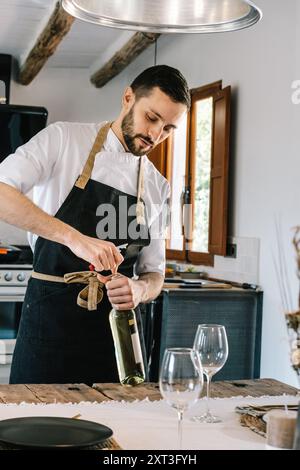 Chef privé dans une cuisine rustique, se concentrant intentionnellement sur l'ouverture d'une bouteille de vin blanc. Il regarde vers le bas le décapsuleur, portant un tablier noir ov Banque D'Images