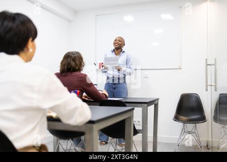 Une femme afro-américaine enthousiaste se tient debout, menant une discussion dans une salle de classe moderne, engageant avec des étudiants adultes multiethniques assis aro Banque D'Images