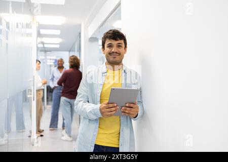 Un jeune homme joyeux en tenue décontractée tient une tablette numérique tout en se tenant debout dans un couloir d'école en arrière-plan, les collègues engagent la conversation Banque D'Images