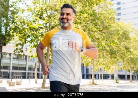 Un Indien, regardant loin, court à travers un parc ensoleillé avec le sourire, portant une chemise sportive jaune et blanche avec une smartwatch noire. Banque D'Images