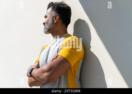 Un Indien portant une chemise de sport jaune et une montre de fitness se tient debout avec les bras croisés, regardant loin contre un mur blanc. Banque D'Images
