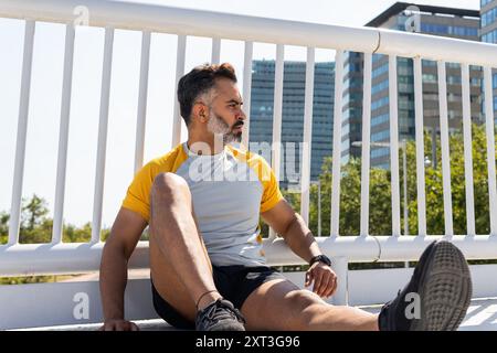 Homme indien en vêtements de sport assis et regardant loin sur une journée d'été ensoleillée, se reposant après une séance d'entraînement, avec des bâtiments de la ville en arrière-plan. Banque D'Images