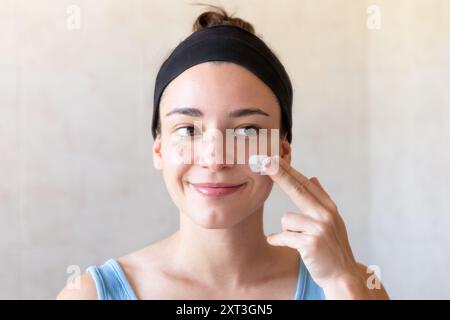 Une jeune femme dans un débardeur bleu, appliquant de la crème hydratante sur son visage, se tient devant un fond neutre, incarnant une routine quotidienne de soins personnels Banque D'Images