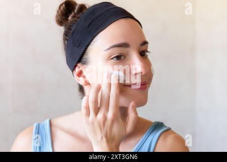 Une jeune femme avec un bandeau sourit tout en appliquant de la crème sur son visage, incarnant un rituel de soin de la peau détendu et sain Banque D'Images