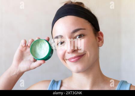 Une jeune femme avec un bandeau sourit alors qu'elle tient un récipient ouvert de masque facial vert, démontrant une routine d'auto-soins dans la salle de bain Banque D'Images