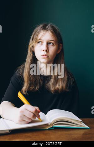 Une étudiante s'assoit à un bureau étudiant à partir d'un manuel ouvert. Regardant loin de la caméra, elle tient un stylo jaune, profondément dans la pensée contre un g sombre Banque D'Images