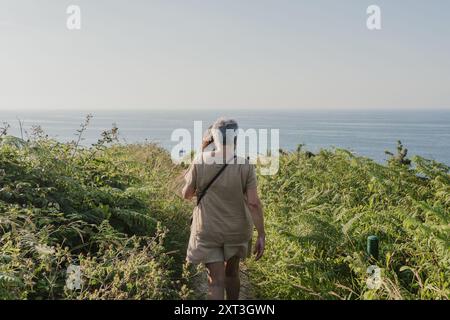 Vue arrière d'une femme âgée marchant paisiblement le long d'un sentier côtier luxuriant. L'océan bleu tranquille s'étend derrière elle sous un ciel dégagé. Banque D'Images