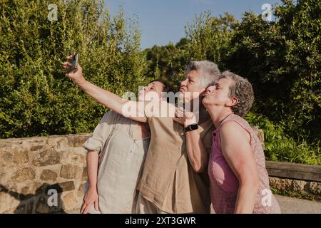 Trois amies âgées, deux femmes partagent un moment joyeux en prenant un selfie avec un téléphone portable dans un cadre de parc luxuriant. Banque D'Images