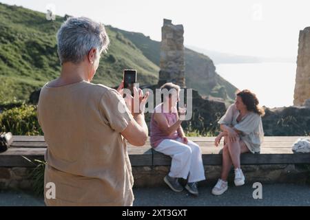 Vue arrière d'une femme âgée capture une photo de ses amis avec un smartphone à un endroit pittoresque. Ses compagnons s'engagent dans la conversation, overlooki Banque D'Images